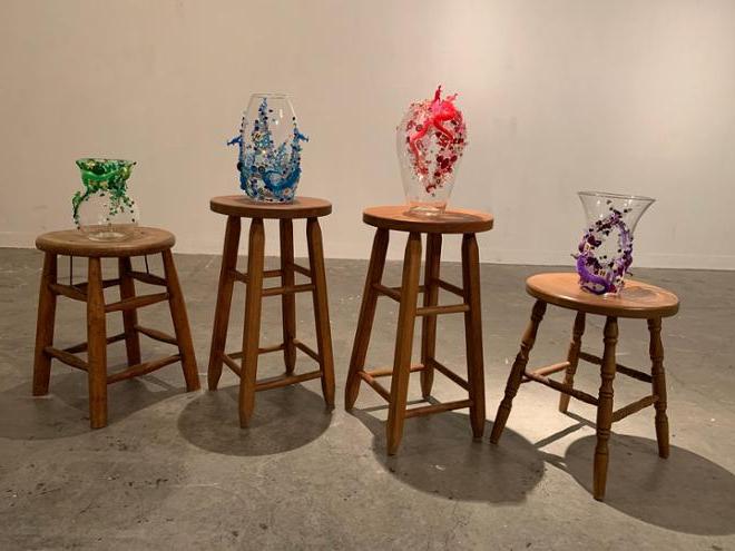 A picture of the same four wooden stools with the vases on them all in a line. In order from left to right the colors are purple, pink, blue, and green.  