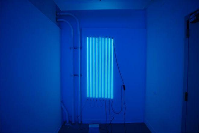 九根透明的氖气水银管，垂直地挂在白色的墙上.  每根管子之间的间距为3英寸，水平间距为2英尺.  从灯管发出的光以一种浅蓝色的光充满了整个空间. 